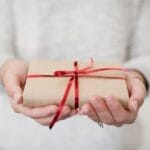 Hands holding a small gift wrapped in brown paper with a red ribbon, symbolizing the simple joy of Christ centered Christmas giving.