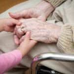 A young child gently holding the hands of an elderly woman sitting in a wheelchair, symbolizing love, care, and multigenerational family support.