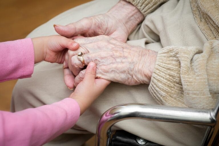 A young child gently holding the hands of an elderly woman sitting in a wheelchair, symbolizing love, care, and multigenerational family support.