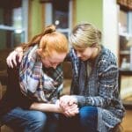 Two women sitting close together on a couch, praying and holding hands with heads bowedโdemonstrating Christ-centered friendship, encouragement, and gospel grace in a moment of vulnerability.