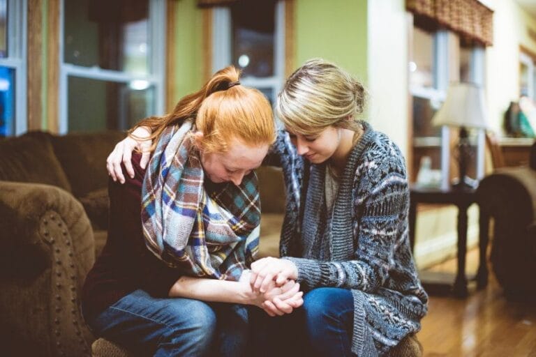 Two women sitting close together on a couch, praying and holding hands with heads bowed—demonstrating Christ-centered friendship, encouragement, and gospel grace in a moment of vulnerability.