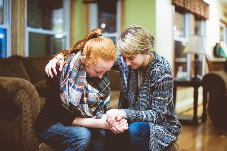 Two women sitting close together on a couch, praying and holding hands with heads bowed—demonstrating Christ-centered friendship, encouragement, and gospel grace in a moment of vulnerability.