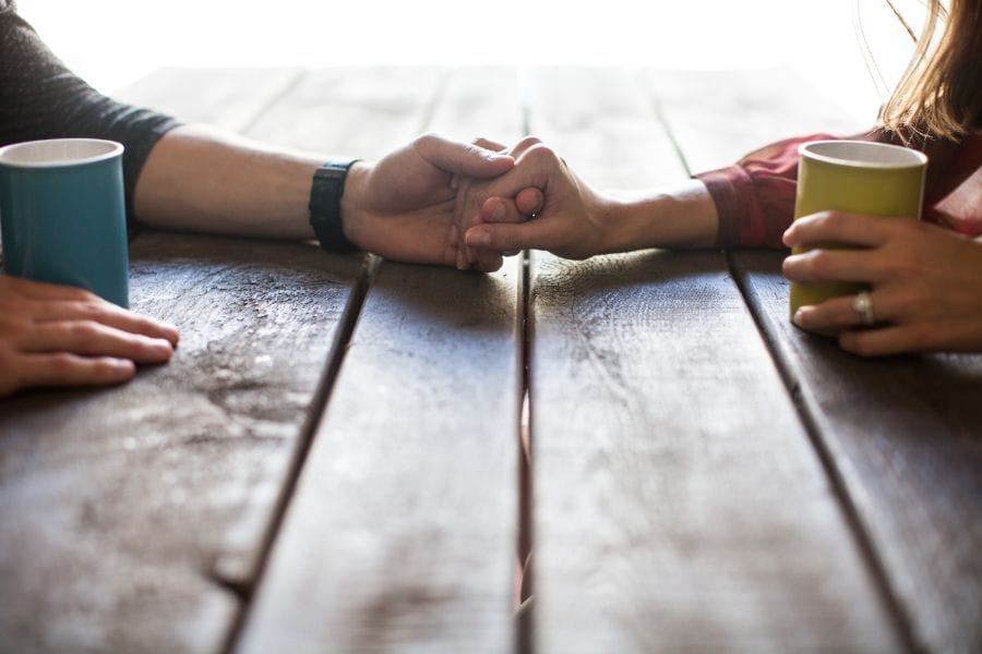 Husband and wife holding hands across a rustic wooden table, each with a coffee mug, symbolizing love, connection, and quiet encouragement in marriage.