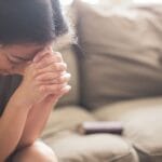 Woman sitting on a couch with head bowed and hands clasped in prayer, with a Bible beside her โ a quiet moment of faithful prayer.