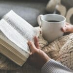 A woman holding an open Bible and a mug of coffee while wrapped in a cozy knit blanket, sitting in a peaceful, homey setting—representing quiet time in God’s Word.