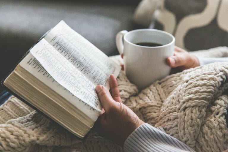 A woman holding an open Bible and a mug of coffee while wrapped in a cozy knit blanket, sitting in a peaceful, homey setting—representing quiet time in God’s Word.