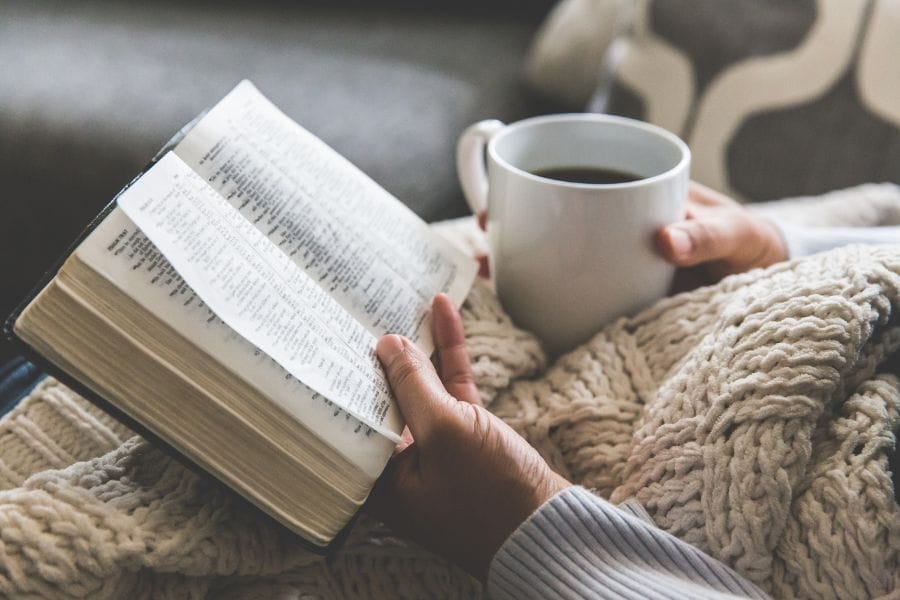 A woman holding an open Bible and a mug of coffee while wrapped in a cozy knit blanket, sitting in a peaceful, homey setting—representing quiet time in God’s Word.