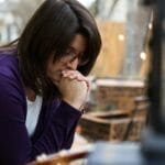 Woman sitting outdoors with hands clasped in prayer, looking thoughtful and reflective. A warm light glows in the foreground, creating a peaceful setting.