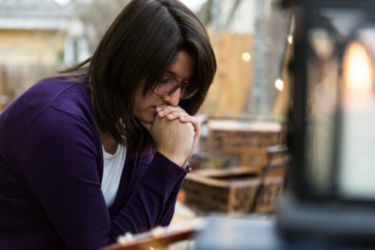 Woman sitting outdoors with hands clasped in prayer, looking thoughtful and reflective. A warm light glows in the foreground, creating a peaceful setting.