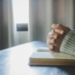 Hands clasped in prayer over an open Bible on a wooden table, with soft natural light coming through a window—symbolizing humility, reflection, and seeking God’s wisdom over self-promotion.