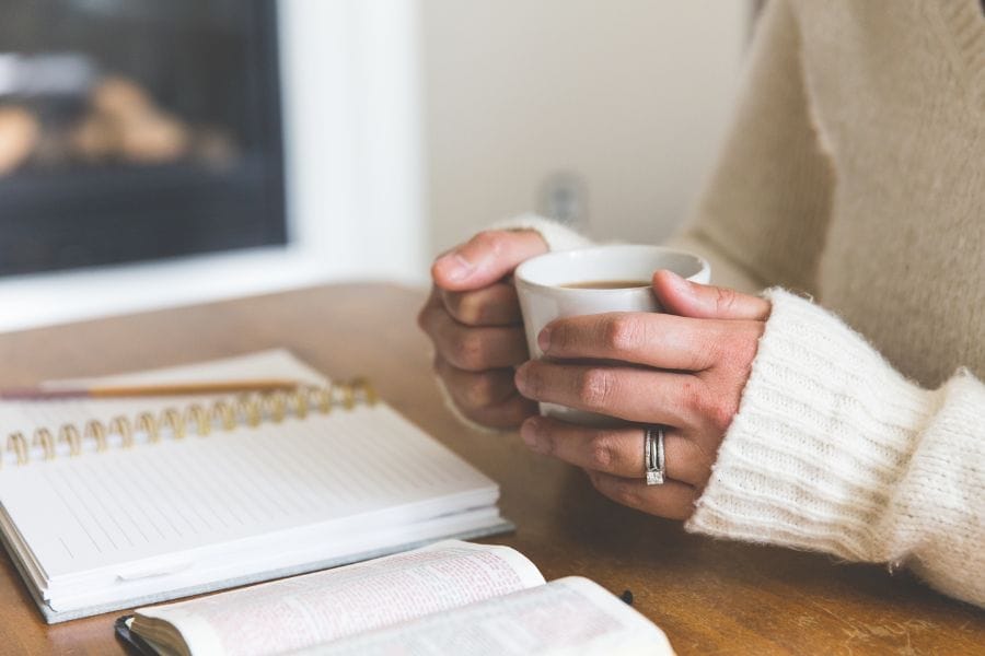 A woman in a cozy cream sweater holding a coffee mug beside an open Bible and journal, symbolizing quiet time with the Lord and simple, faithful living.