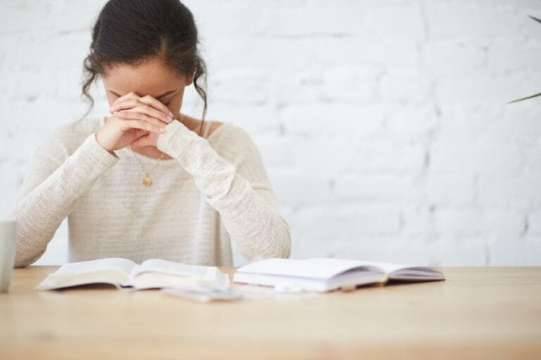 Woman praying with head bowed over an open Bible and journal at a table, symbolizing quiet time with the Lord and heart-focused prayer.