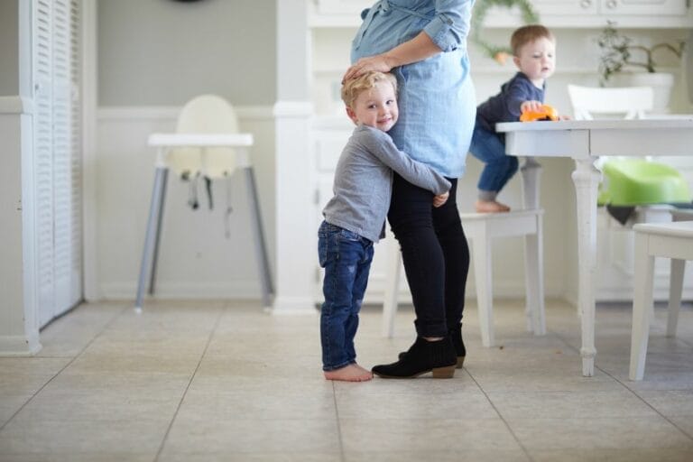 Young boy hugging his mother in a bright, peaceful kitchen while another child plays at the table—capturing a life-giving moment of love and connection in everyday homemaking.