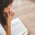 A woman with folded hands prays over an open Bible, symbolizing a quiet moment of reflection and trust in God during personal prayer.