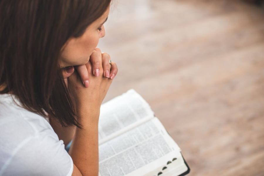 A woman with folded hands prays over an open Bible, symbolizing a quiet moment of reflection and trust in God during personal prayer.