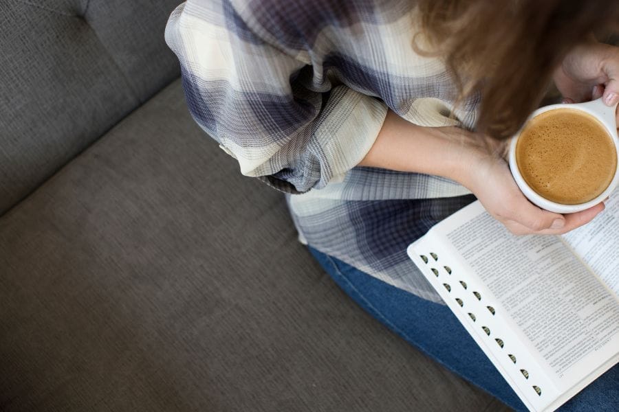 Woman sitting on a gray couch holding a cup of coffee and an open Bible, ready for quiet time with the Lord.