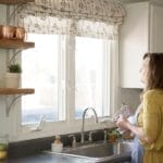 A woman washing dishes at her kitchen sink and looking out the window, reflecting the quiet, everyday ministry that happens in the home.