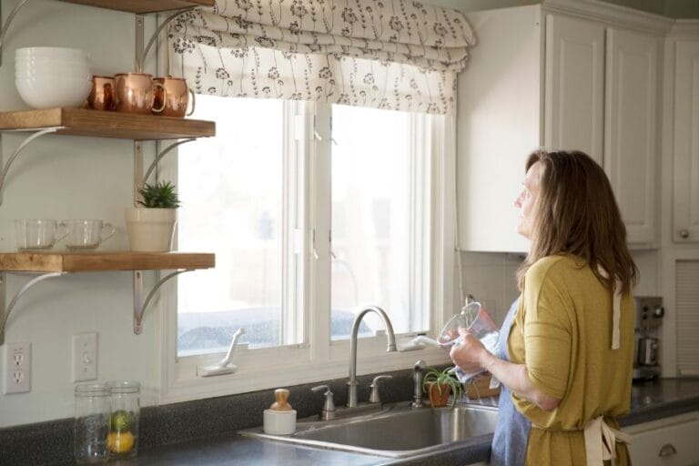 A woman washing dishes at her kitchen sink and looking out the window, reflecting the quiet, everyday ministry that happens in the home.