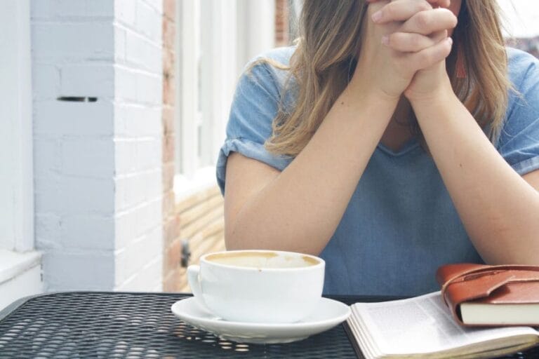 A woman sitting at a café table with a Bible and journal open, hands clasped in prayer beside a cup of coffee, reflecting on God’s Word and the sin of selfishness.
