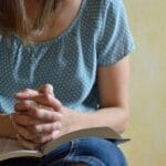 Woman sitting in prayer with hands folded over an open Bible, a quiet moment of trust and reflection.