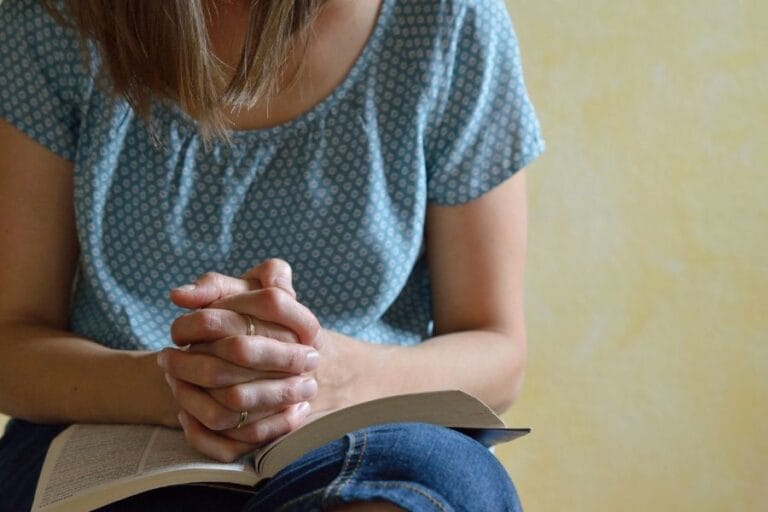 Woman sitting in prayer with hands folded over an open Bible, a quiet moment of trust and reflection.