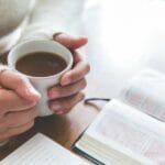 A woman holding a cup of coffee next to an open Bible and journal, reflecting on God’s faithfulness and trusting His work in the lives of others.
