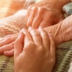 Close-up of a younger hand gently resting on an elderly woman’s hand, both wrapped in a warm blanket—symbolizing love, caregiving, and gospel-centered end-of-life comfort.