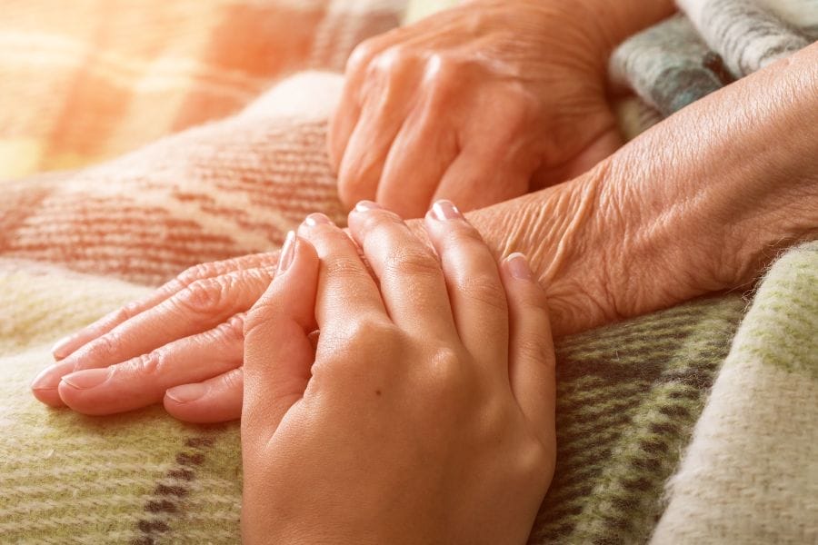 Close-up of a younger hand gently resting on an elderly woman’s hand, both wrapped in a warm blanket—symbolizing love, caregiving, and gospel-centered end-of-life comfort.