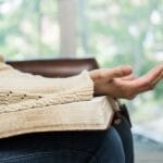 A woman sitting with an open Bible on her lap, wearing a cream sweater, and holding one hand open in a posture of prayer or surrender. Sunlight filters through a window in the background.