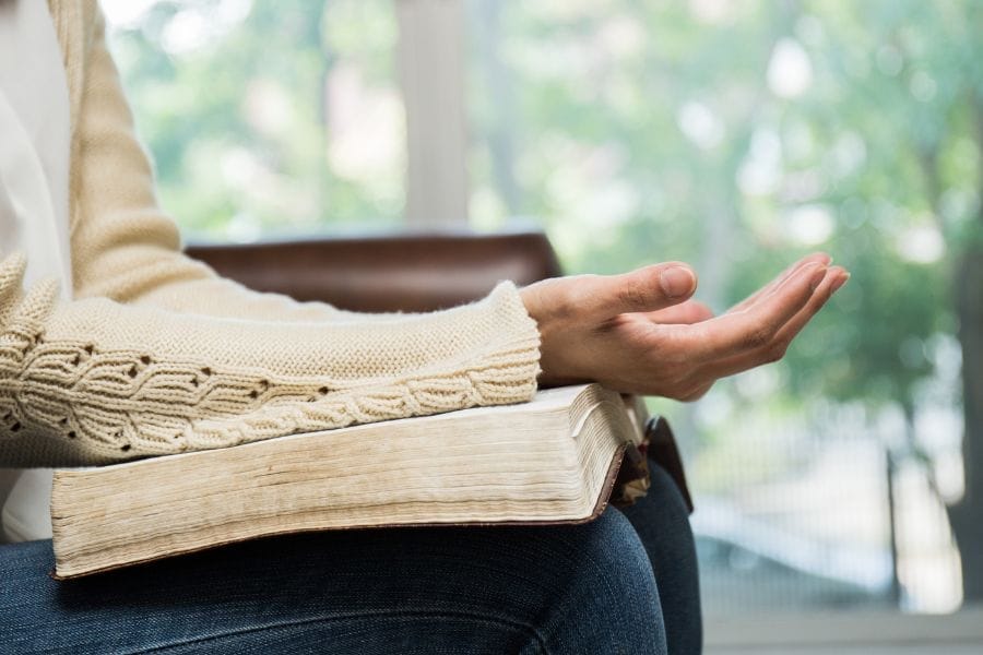 A woman sitting with an open Bible on her lap, wearing a cream sweater, and holding one hand open in a posture of prayer or surrender. Sunlight filters through a window in the background.