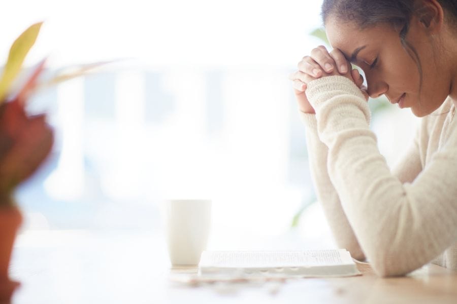 Woman praying beside an open Bible at a table in soft morning light